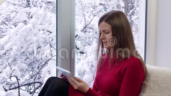 穿红裙子的漂亮女孩坐在窗台上用平板电脑冬天在外面视频的预览图