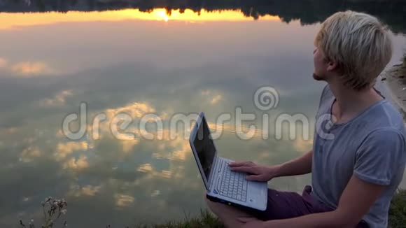 在斯隆莫的夕阳下创意人用笔记本电脑在河岸上坐下视频的预览图