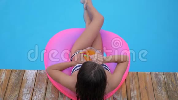 夏天休息的女孩穿上泳衣躺在靠近游泳池的粉红色充气圈的露台上喝着五颜六色的鸡尾酒视频的预览图