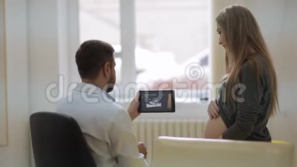 一位身穿白色医用晨衣的医生在平板电脑上展示了一位孕妇的超声波照片视频的预览图