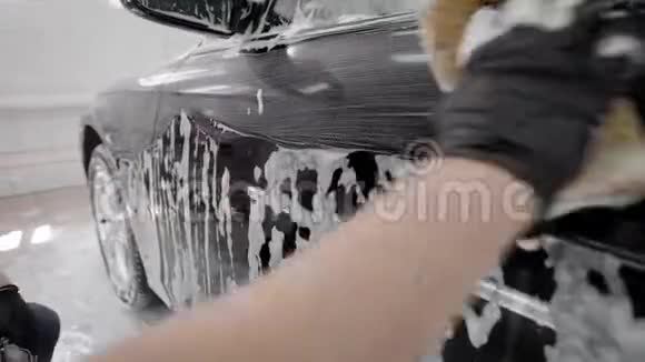 洗衣工在洗车时用海绵摩擦黑色汽车表面用肥皂泡沫擦拭戴橡胶手套视频的预览图