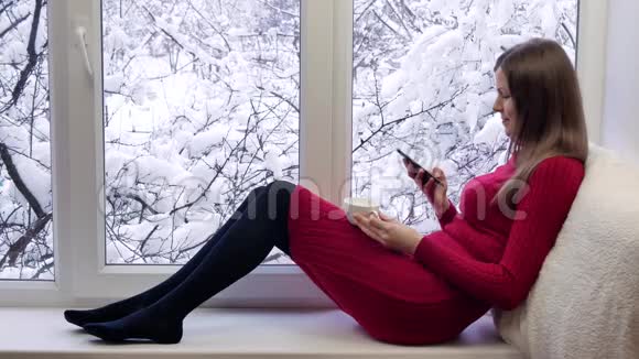 穿着红裙子的漂亮女孩坐在窗台上喝咖啡用智能手机冬天在外面视频的预览图