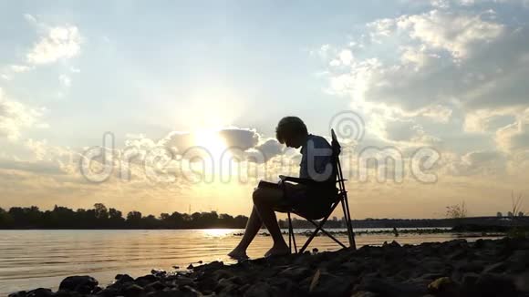 聪明的人坐在河岸上写作把脚放在水里视频的预览图