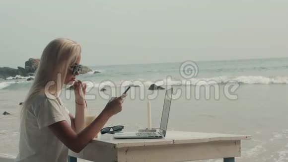 有笔记本电脑和新鲜果汁的女人坐在海滩咖啡馆里在智能手机上聊天海景海浪视频的预览图