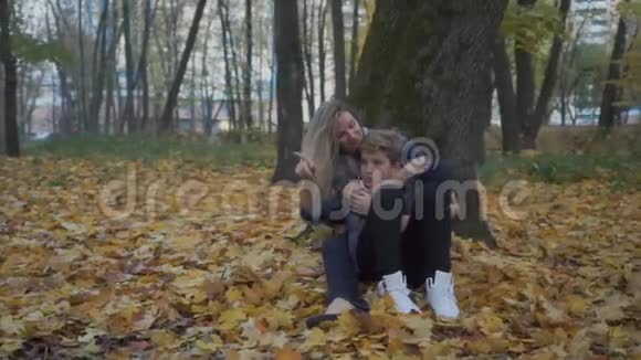 儿子和母亲坐在秋天公园的地上放松视频的预览图