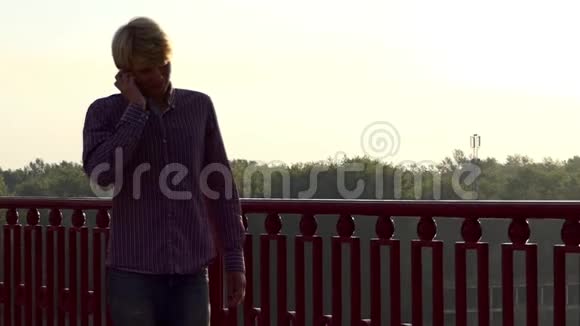年轻人沿着大桥走在斯隆莫的日落处由一个移动电话交谈视频的预览图
