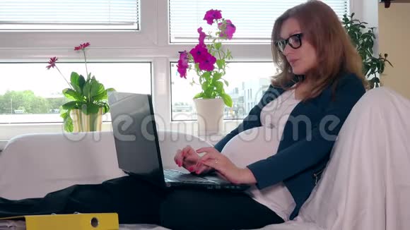 一位怀孕的女企业家她拿着笔记本电脑坐在沙发上视频的预览图