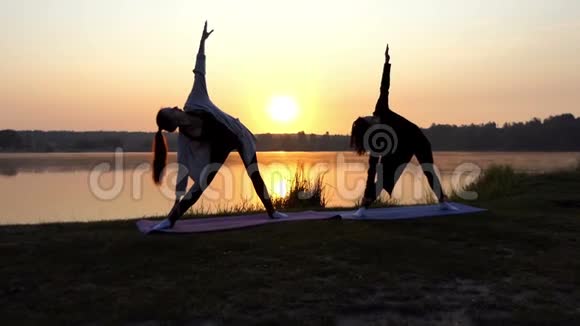 两个女人站在瑜伽垫上在日落的湖岸上弯腰视频的预览图