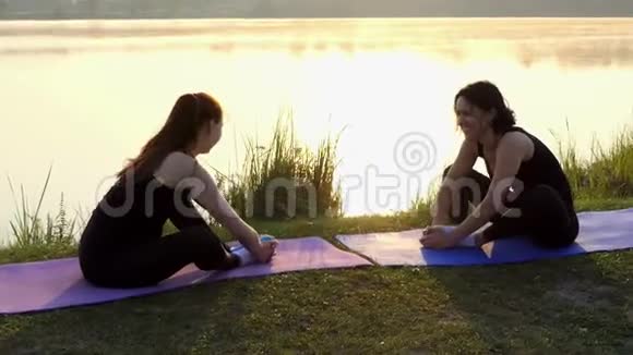 两个女人坐在垫子上向前弯曲在日落时双脚并拢视频的预览图