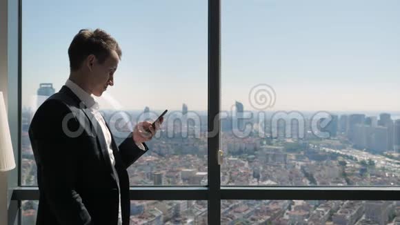 在办公室的年轻商人正在窗口附近浏览电话可以看到全景城市景色视频的预览图