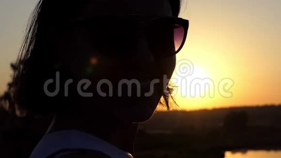 一个微笑的年轻女子在缓慢的动作中愉快地透过太阳镜看日落视频的预览图