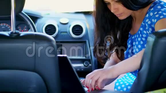 美丽的黑发女人穿着蓝色连衣裙坐在车里正在笔记本电脑上工作在社交网络上交流使用视频的预览图