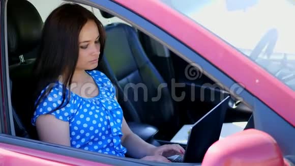 美丽的黑发女人穿着蓝色连衣裙坐在车里正在笔记本电脑上工作在社交网络上交流使用视频的预览图