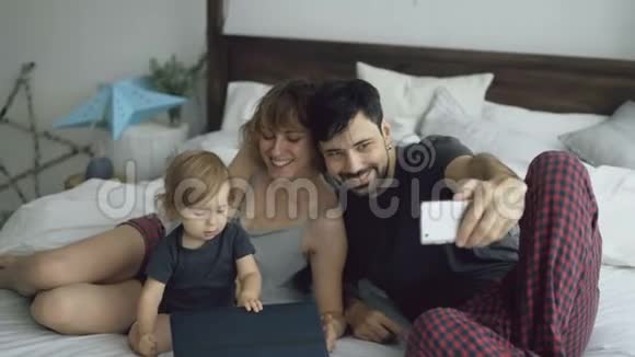 年轻的一家人带着可爱的小女孩坐在家里的智能手机摄像头上自拍视频的预览图