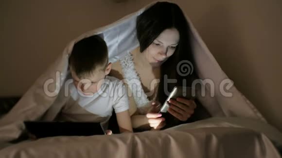 年轻漂亮的妈妈在手机上浏览互联网而她的儿子在平板电脑上看卡通视频的预览图