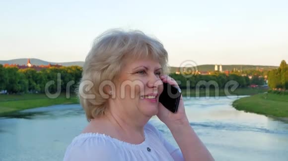 老妇人站在河上的桥上用智能手机说话视频的预览图