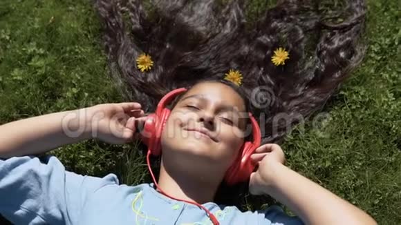 漂亮的长发女孩躺在公园的草地上听着耳机上的音乐慢动作高清高清视频的预览图