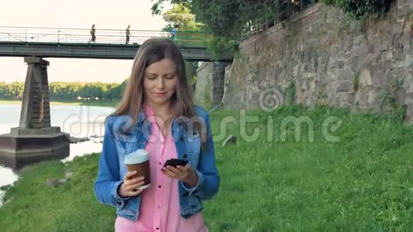漂亮的年轻女孩使用智能手机和喝咖啡沿着海滨散步灞上野石桥和老墙视频的预览图