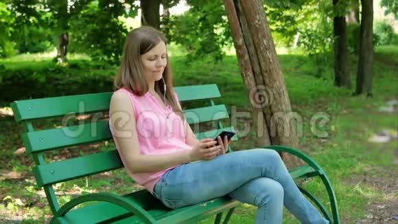 年轻女子坐在公园的长凳上用智能手机带耳机使用智能手机的漂亮女孩视频的预览图