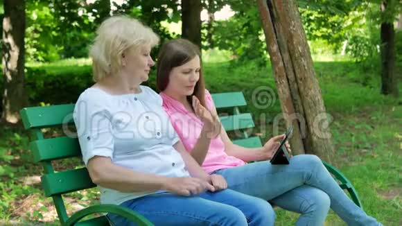 坐在公园的长凳上通过电脑平板电脑打视频电话的老年妇女和年轻妇女成年母亲视频的预览图