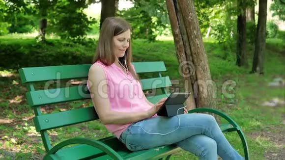 年轻女子坐在公园的长凳上用带耳机的平板电脑使用平板电脑的美女视频的预览图