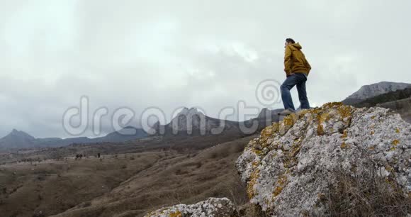 一个穿着黄色夹克衫的人站在一块石头上站在雾中白雪覆盖的山峰背景下视频的预览图