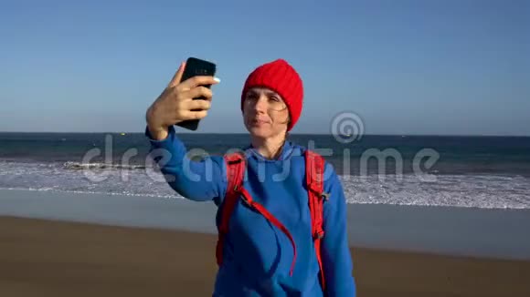 活跃的徒步旅行者女人走在海滩上在她的智能手机上自我拍照带背包的白人年轻女子视频的预览图