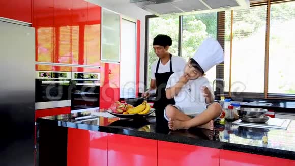 泰国私人厨师烹饪他的小儿子在厨师的帽子附近坐在桌子上在一个现代风格的家庭厨房孩子在吃东西视频的预览图