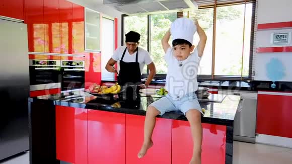 泰国私人厨师烹饪他的小儿子在厨师的帽子附近坐在桌子上在一个现代风格的家庭厨房孩子在吃东西视频的预览图
