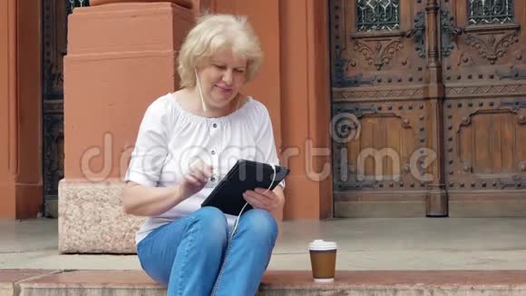 老妇人坐在台阶上用带耳机的平板电脑喝咖啡背景中的老式建筑视频的预览图