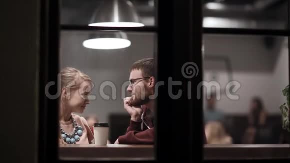 通过窗口查看年轻夫妇坐在咖啡馆里喝咖啡聊天晚上一起玩视频的预览图