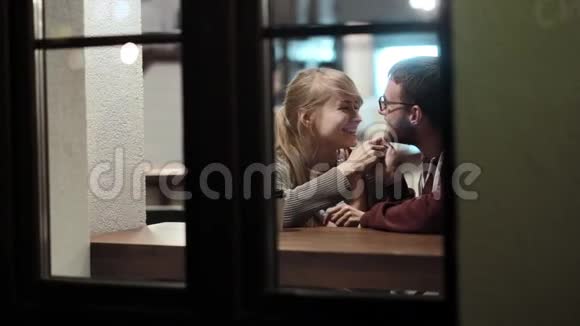 通过窗口查看快乐的年轻夫妇晚上在咖啡馆里坐在桌子旁接吻视频的预览图