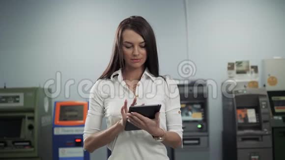 一幅美丽的中年妇女在ATM机前银行办公室的画像手里拿着数字平板电脑和视频的预览图