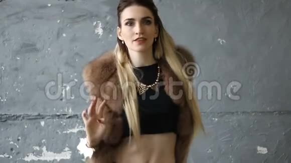 年轻迷人的金发女郎穿着人造毛皮敞开着前背心和时髦的黑色上衣在灰色背景上跳舞视频的预览图