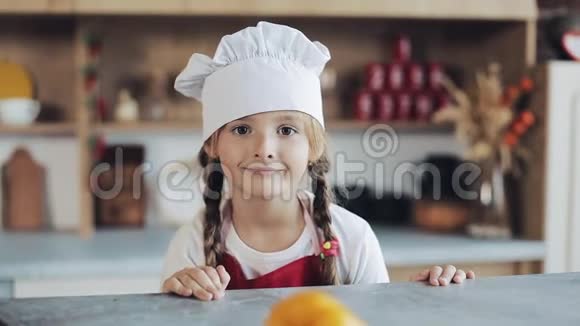 厨房里一个打扮成职业厨师的可爱小女孩看着镜头微笑的画像视频的预览图