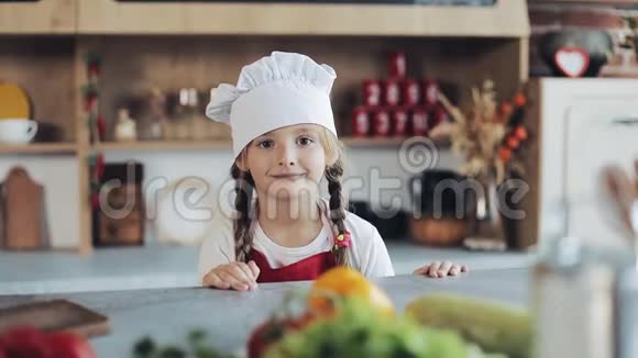 厨房里一个打扮成职业厨师的可爱小女孩看着镜头微笑的画像视频的预览图