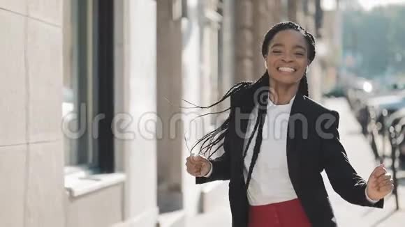 年轻快乐的非裔美国女商人戴着耳机在城市的大街上走着一边听着音乐视频的预览图