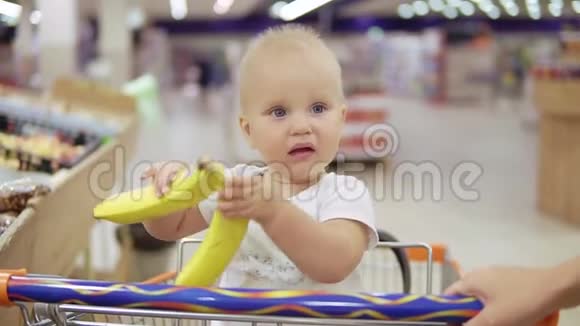 可爱的小孩正在帮助她的母亲在超市里挑选香蕉而坐在购物车里手里拿着香蕉视频的预览图