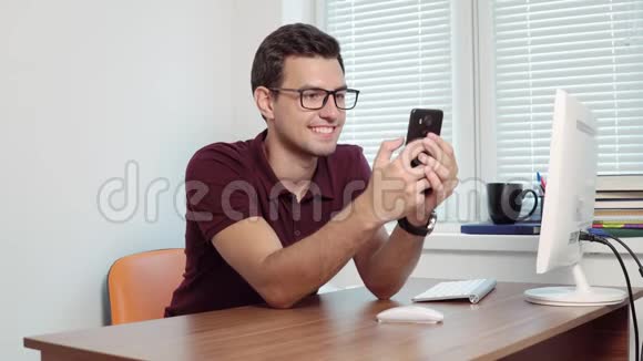男人看着手机的屏幕微笑着在办公室的坐桌上发短信英俊的年轻商人视频的预览图