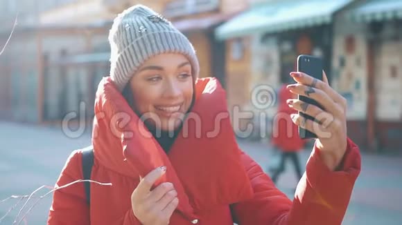 穿着红色冬季夹克的迷人女孩在明亮的冬天站在冬天的街道上在手机上自拍视频的预览图