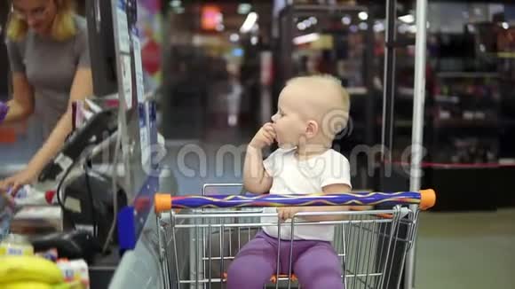 年轻的妈妈在超市的传送带上拿出手推车的产品而她可爱的小宝宝在吃东西视频的预览图