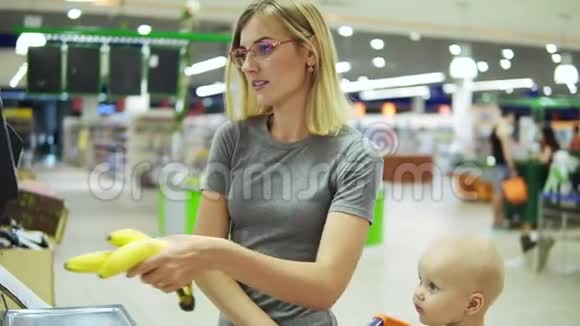 年轻的母亲在超市里用自助式电子秤称香蕉而她可爱的孩子则坐在超市里视频的预览图