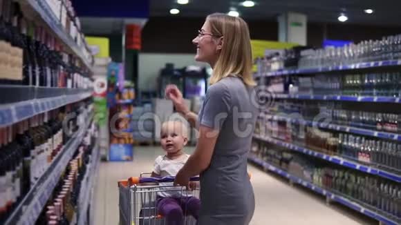 漂亮的年轻妈妈正在超市的饮料部挑选一瓶酒而她的小宝贝视频的预览图