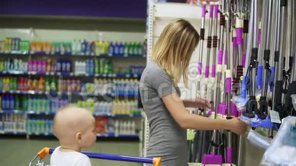 漂亮的女人在超市里选择地板拖把而她的孩子坐在购物车里家庭视频的预览图