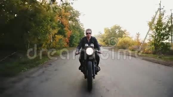 在阳光明媚的柏油路上骑摩托车一个戴着太阳镜和皮夹克的时尚酷青年的正面景色视频的预览图