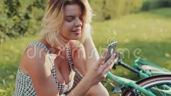 漂亮的金发女郎穿着时髦漂亮的连衣裙坐在城市公园的绿草上骑着老式自行车视频的预览图