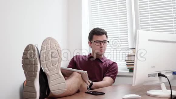 戴眼镜的年轻工人用双腿握着键盘打字用电脑屏幕看着办公室里的电脑视频的预览图