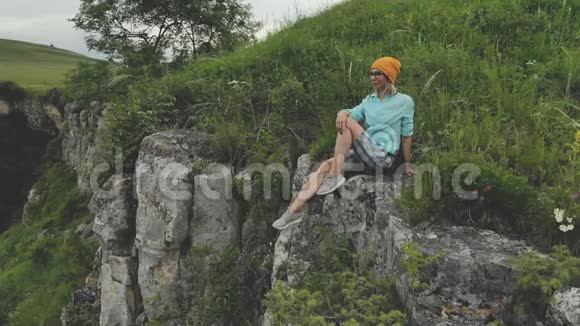 年轻的时髦女人坐在高高的岩石墙上的边缘空中观景无人机缓慢地向后飞行视频的预览图