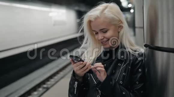 年轻的金发旅行女人手里拿着手机在火车站台上等着公共交通视频的预览图