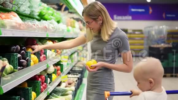 年轻的母亲正在选择黄色的西红柿和其他蔬菜而她的孩子正坐在一辆购物车里视频的预览图